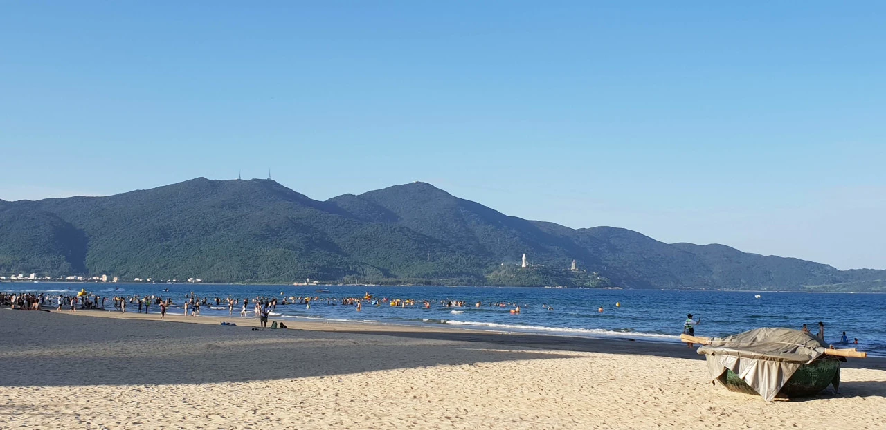 Plage de sable jaune à Da Nang au Vietnam, avec la mer et des montagnes verdoyantes en arrière-plan. Un bateau-panier rond traditionnel (Thung Chai) est visible sur le sable à droite.