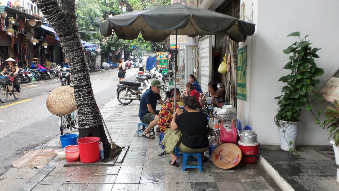 Scène de vie locale à Hanoï : des voyageurs et des habitants installés sur des petits tabourets en plastique pour manger sur le trottoir, entourés de scooters stationnés et de vélos circulant dans la rue.